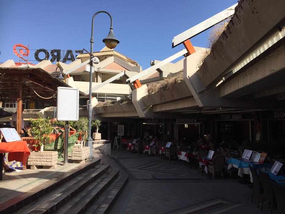 Eine Straße, die in einen Essbereich mit Tischen, Stühlen, Büchern und Gegenständen verwandelt wird, umgeben von Gebäuden, Straßenmöbeln und Grünflächen unter einem klaren Himmel.