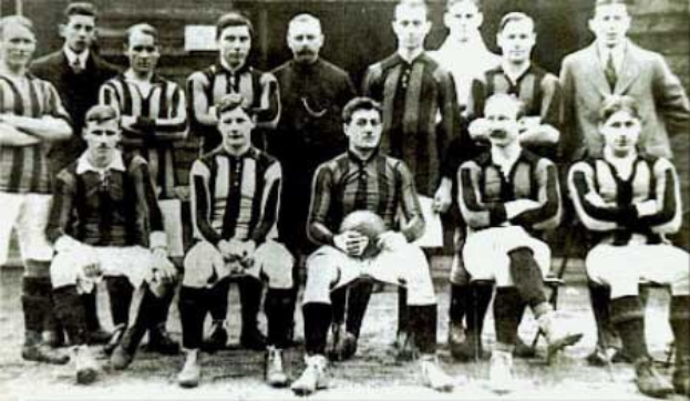 Schwarzes und weißes Foto einer Fußballmannschaft, einige stehen und einige sitzen, vor einer Wand mit dem Text "Ballymena Football Club" unten.