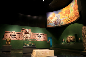 Ausstellung verschiedener Artefakte im Anthropologischen Museum von Mexiko-Stadt, darunter Skulpturen, Tischgegenstände, eine Texttafel, eine Wandmalerei und Deckenbeleuchtung.