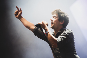 Ein Mann in einem schwarzen Hemd und Jeans singt leidenschaftlich in ein Mikrofon auf einer hell beleuchteten Bühne, mit einem engagierten Publikum, das im Hintergrund klatscht und jubelt.