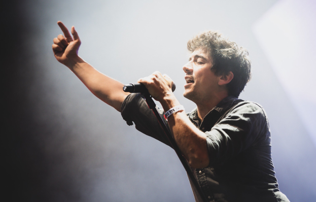 Ein Mann in einem schwarzen Hemd und Jeans singt leidenschaftlich in ein Mikrofon auf einer hell beleuchteten Bühne, mit einem engagierten Publikum, das im Hintergrund klatscht und jubelt.