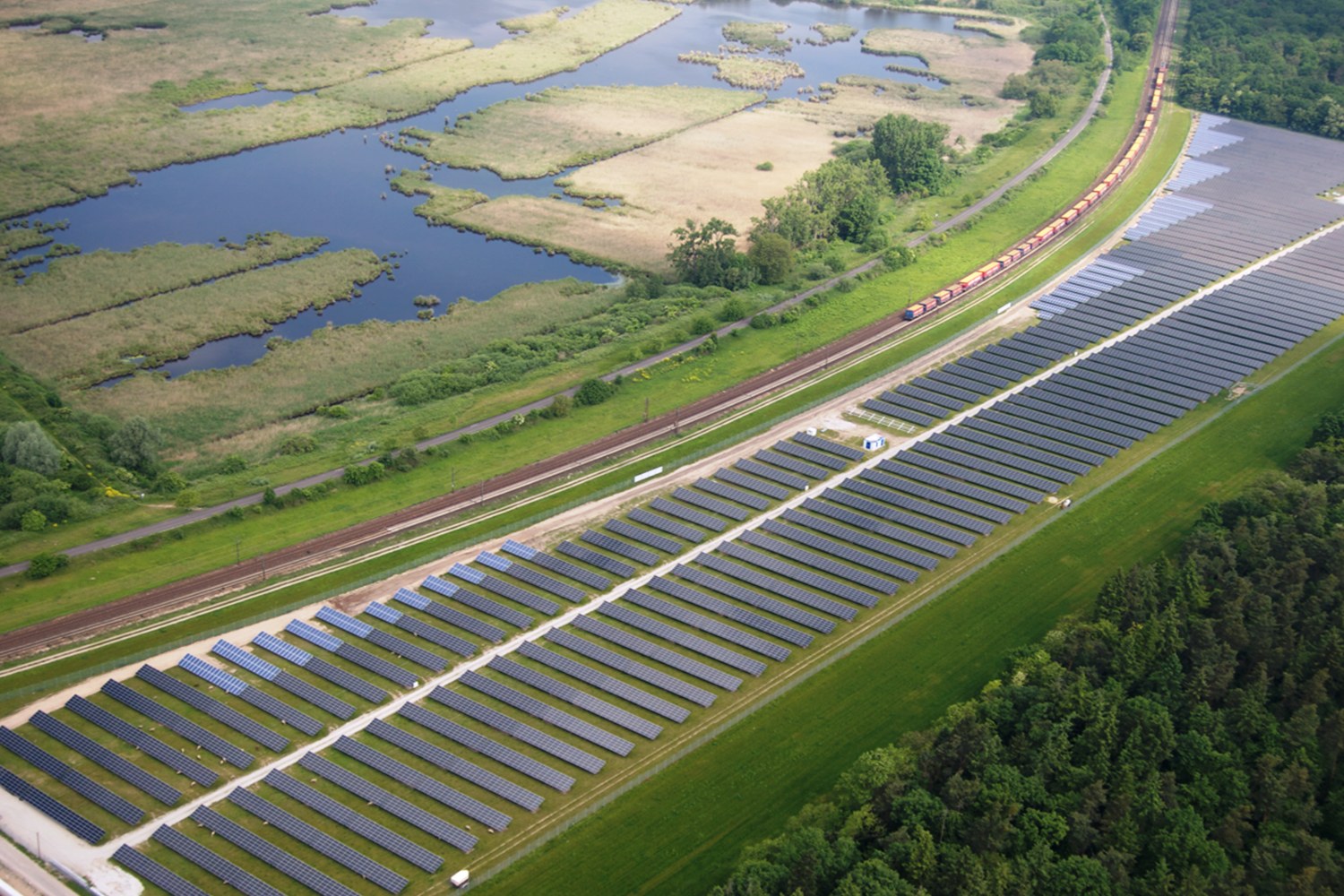 Luftaufnahme einer großen Solaranlage mit Solarpanelen in einem Feld, umgeben von Bäumen, Gras, Pflanzen und Wasser, mit einem Zug auf einer nahen Eisenbahnschiene.