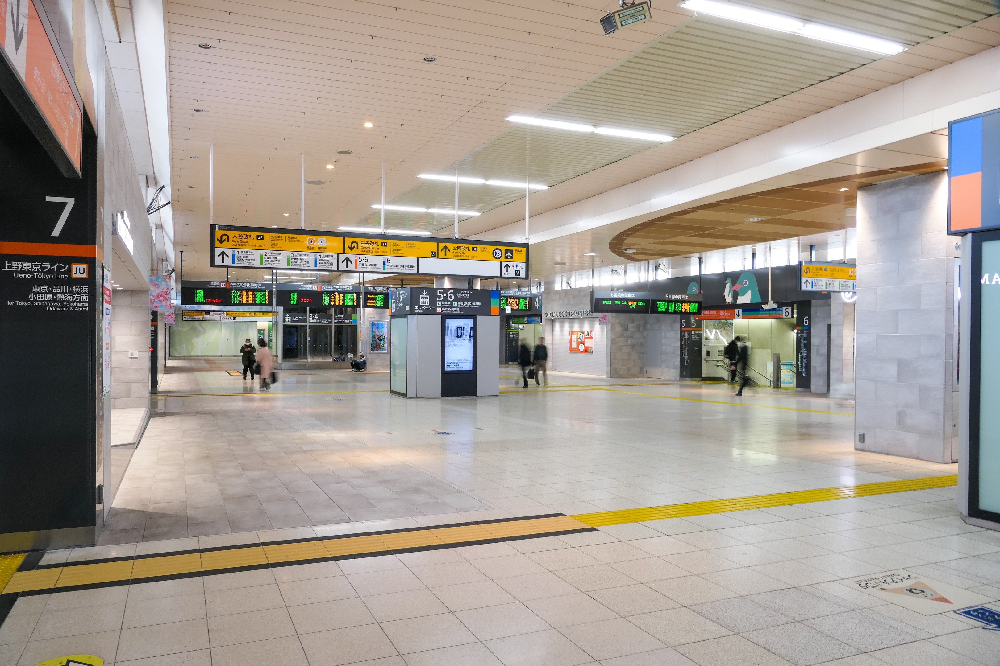 Leerer Bahnhof mit Menschen, Anzeigetafeln an den Wänden, Deckenleuchten und tragenden Säulen.