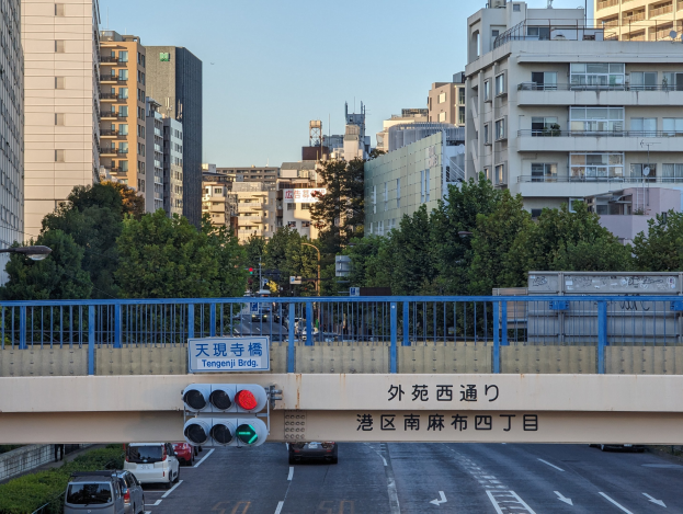 Eine Straße in einer Stadt mit fahrenden Autos, eine Brücke mit Geländern und ein Schild, Verkehrsampeln, Laternen, Bäume und Gebäude mit Fenstern unter einem klaren Himmel.