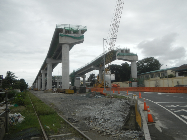 Baustelle mit einer Brücke im Hintergrund, eine Straße mit Absperrgittern auf der rechten Seite, Steine und Gras am Boden, eine Bahnschiene auf der linken Seite, Bäume und Gebäude auf beiden Seiten der Straße und ein bewölkter Himmel.