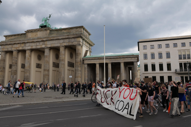 Gruppe von Menschen zu Fuß und mit dem Fahrrad auf einer Straße vor dem Brandenburger Tor, die eine Fahne halten, mit Laternenmasten und einem bewölkten Himmel im Hintergrund.
