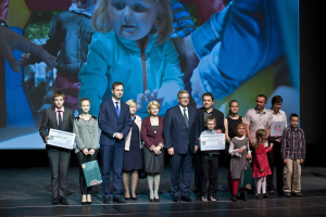 Gruppe von Menschen auf einer Bühne, die Tüten und Urkunden halten, mit einem Bildschirm im Hintergrund, der ein Kind und andere zeigt.