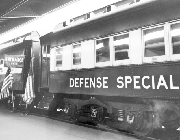 Schwarzes und weißes Foto eines Zuges mit der Aufschrift "Defense Special", der auf Gleisen geparkt ist, mit zwei Fahnen links und einer Decke oben.