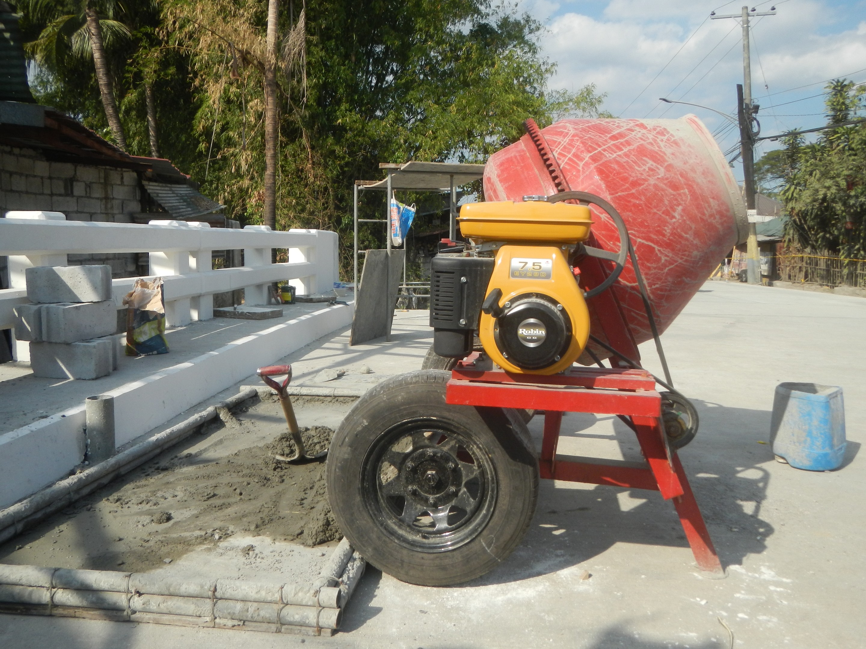 Betonmischer auf einem Zementlaster umgeben von Baustoffen und einem Haus mit Bäumen unter einem bewölkten Himmel.