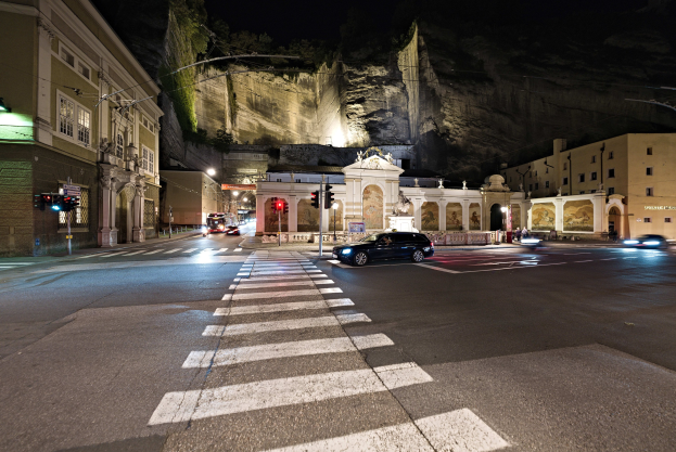 Stadtstraße bei Nacht mit fahrenden Autos, Straßenlaternen, Ampeln, beleuchteten Fenstern, Laternenpfählen, einem Hügel im Hintergrund und einer Zebrastreifenkreuzung unten.