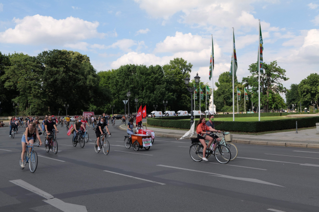 Eine Gruppe von Menschen, die auf Fahrrädern eine von Bäumen gesäumte Straße mit Laternen- und Fahnenmasten entlangfahren, während der Pariser Fahrradtour 2016, mit bewölktem Himmel und ein paar Fahrzeugen im Hintergrund.