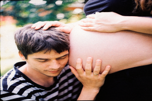 Ein Mann und eine Frau stehen Seite an Seite, der Mann berührt zärtlich den Bauch der Frau, mit einem unscharfen Hintergrund.