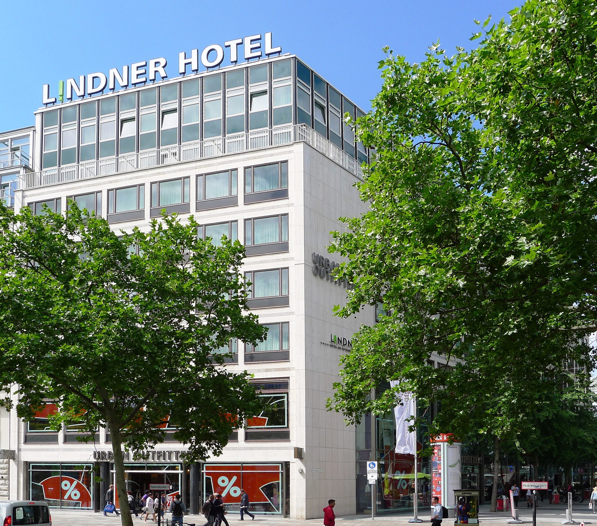 Großes modernes Gebäude mit Glasfenstern und Text auf dem Dach, beschriftet mit "Lindner Hotel", umgeben von Bäumen und einer belebten Straße mit Fußgängern und Fahrzeugen.