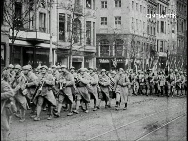 Schwarzes und weißes Foto von Soldaten, die während des Ersten Weltkriegs eine Straße in Berlin entlangmarschieren, mit Mützen und Gewehren, sowie Bäumen und Gebäuden am Straßenrand und einem Pfahl im Hintergrund.