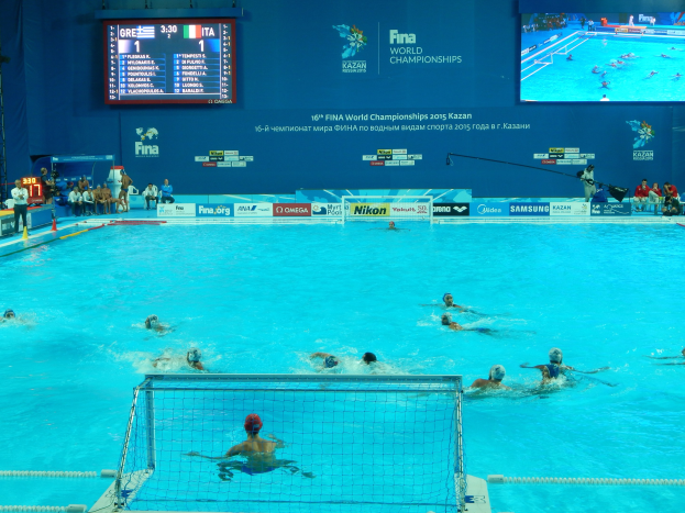 Eine Gruppe von Menschen, die Wasserball in einem großen Pool mit einem Netz in der Mitte spielen, Zuschauer sitzen links und eine Wand im Hintergrund zeigt Text und Logos für die FINA-Weltmeisterschaften 2016 in Kazan.