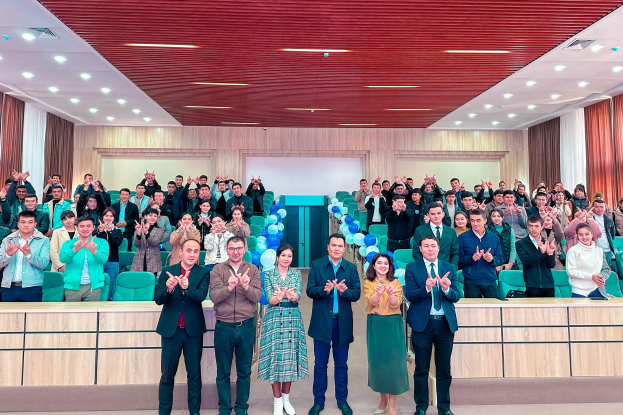 Eine Gruppe von Menschen steht vor einem Publikum, einige klatschen, in einem Raum mit Stühlen, Tischen, Luftballons, Vorhängen und Deckenleuchten, feiernd die Eröffnung einer neuen Schule.