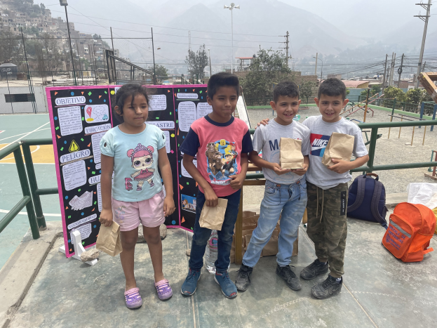 Gruppe von Kindern auf einem Basketballfeld mit Papiertüten in den H├Ąnden, mit Tüten und Gegenst├Ąnden rechts und einer Tafel mit Text links, vor einem Hintergrund aus Zaun, Pfosten, Stra├čenlaternen, B├Ąumen, Geb├Ąuden, H├Ęllen und Himmel.