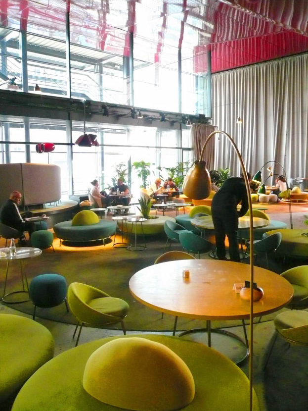 Ein geräumiges Hotel-Foyer mit grünen Stühlen, Tischen, Sofas, stehenden Menschen, Pflanzen, einer Stehlampe, Vorhängen und Deckenleuchten.