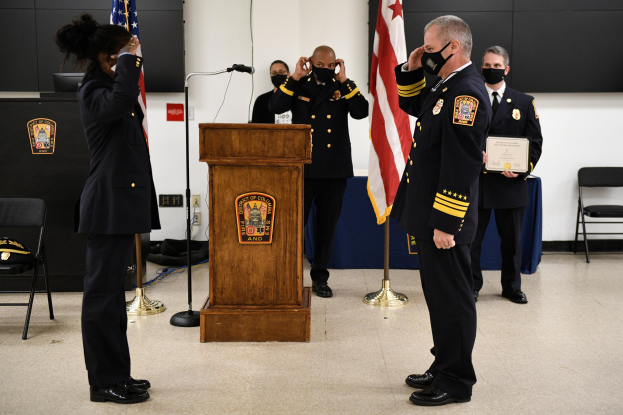 Gruppe uniformierter Personen in Masken vor einem Podium mit Mikrofon, umgeben von Fahnen, Stühlen, Tischen und einer Wand während einer Vereidigung eines neuen Feuerwehrchefs.