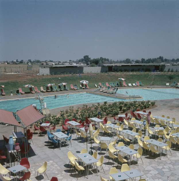 Ein Freibad umgeben von Tischen, Stühlen, Blumenpflanzen, Sonnenschirmen, Pfosten, einem Zaun, einer Wand, einer Baumgruppe, einigen Gebäuden, unter einem bewölkten Himmel.