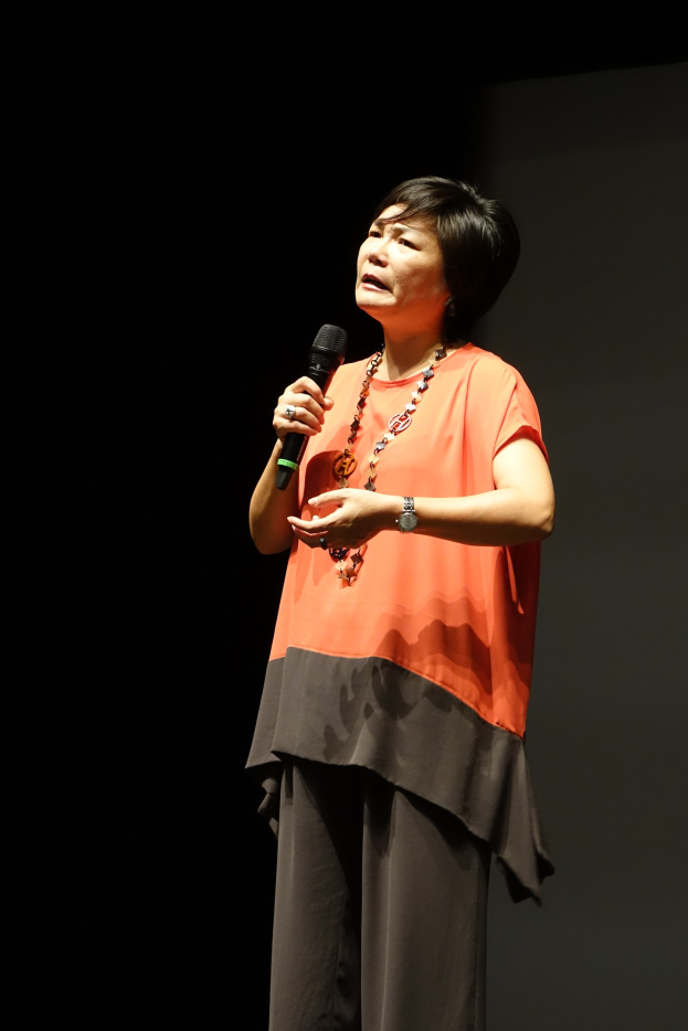 Frau auf einer hell erleuchteten Bühne mit einem Mikrofon in der Hand, die eine weiße Bluse und einen schwarzen Rock trägt und ihr Haar zu einem Dutt gebunden hat und einen selbstbewussten Ausdruck hat.