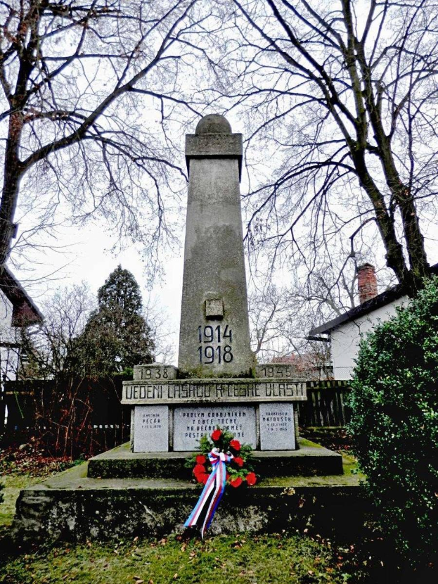 Ein Weltkriegsgedenkmal in einem Friedhof, geschmückt mit einem Kränzchen, umgeben von Gras und trockenen Blättern, mit Pflanzen, Bäumen, Häusern und Himmel im Hintergrund.