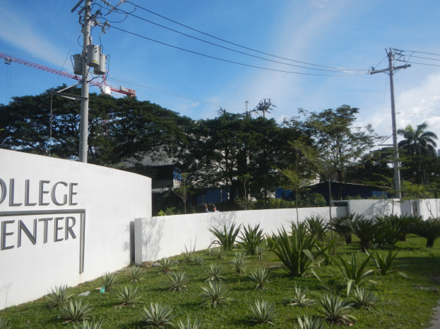 College Center-Eingangsschild an einer Gebäudewand umgeben von Grün, einem Weg, Strommasten, Bäumen, anderen Gebäuden, einem Kran und einem bewölkten Himmel.