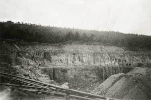 Ein Schwarz-Weiß-Foto einer Baustelle mit einer Brücke im Hintergrund, umgeben von Bäumen und einem klaren Himmel, mit Eisenbahnschienen und Steinen unten.