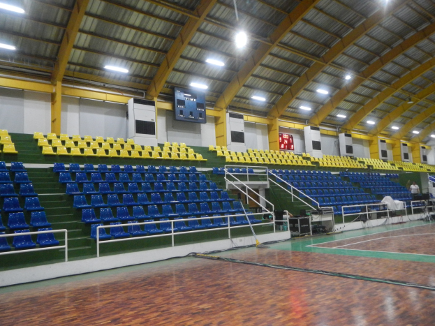 Große Sporthalle mit Holzfußboden, blauen und gelben Sitzplätzen, Geländern, Treppen mit Geländern, Wandbildschirmen, Deckenleuchten und einigen Menschen.