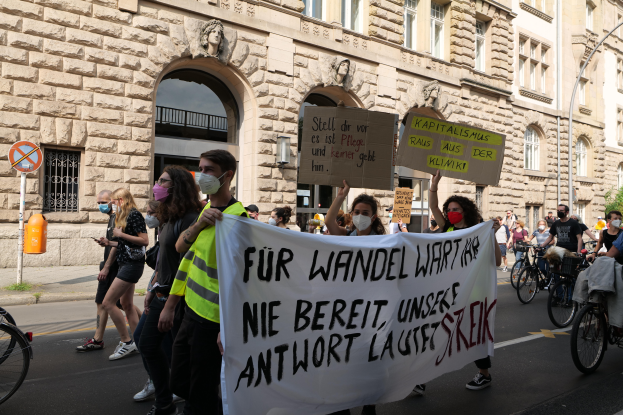 Eine Gruppe von Menschen marschiert auf einer Straße in Berlin, hölt Plakate und Banner, einige fahren Fahrräder, vor einem Gebäude mit Öffnungen, überdächerungen, Pfeilern, Skulpturen und Bäumen.