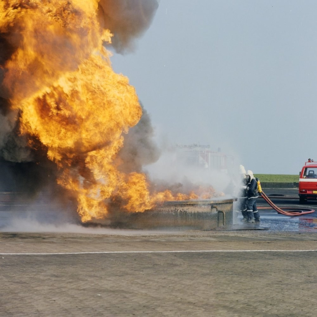 Feuerwehrfahrzeug von Flammen umgeben auf der Seite einer Straße, mit zwei Helden mit Helmen und Rohren und einem Fahrzeug im Hintergrund unter einem Himmel.