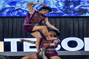 Zwei Performer in violetten Kleidern führen Akrobatik auf der Bühne aus, während das Publikum unten zusieht.