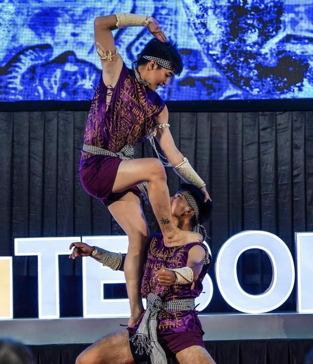 Zwei Performer in violetten Kleidern führen Akrobatik auf der Bühne aus, während das Publikum unten zusieht.