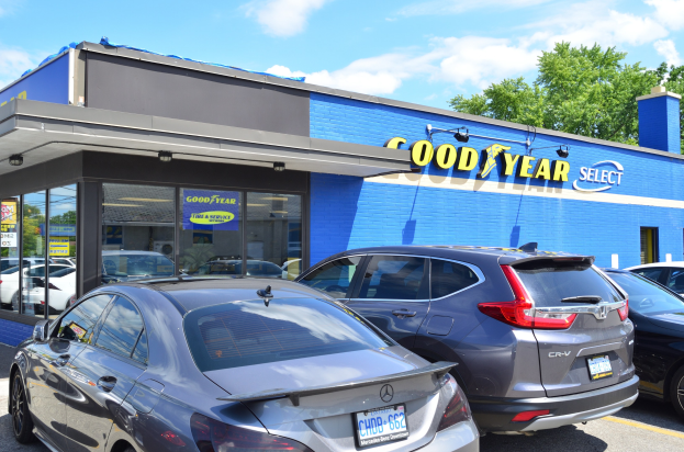 Außenansicht eines Goodyear Select Autohauses mit parkenden Fahrzeugen, sichtbarer Beschilderung, Bäumen im Hintergrund und einem bewölkten Himmel.