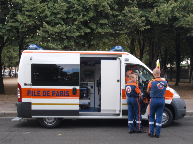 Ambulanz auf dem Seitenstreifen geparkt mit drei Personen davor, Bäume, Gebäude und andere Fahrzeuge im Hintergrund.