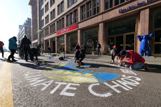 Menschen sitzen vor einem Gebäude mit Fenstern und Namensschildern auf dem Boden, umgeben von Flaschen und anderen Gegenständen, während sie an einer Klimaprotestaktion in Berlin teilnehmen, die von Bäumen und einem klaren blauen Himmel umgeben ist.