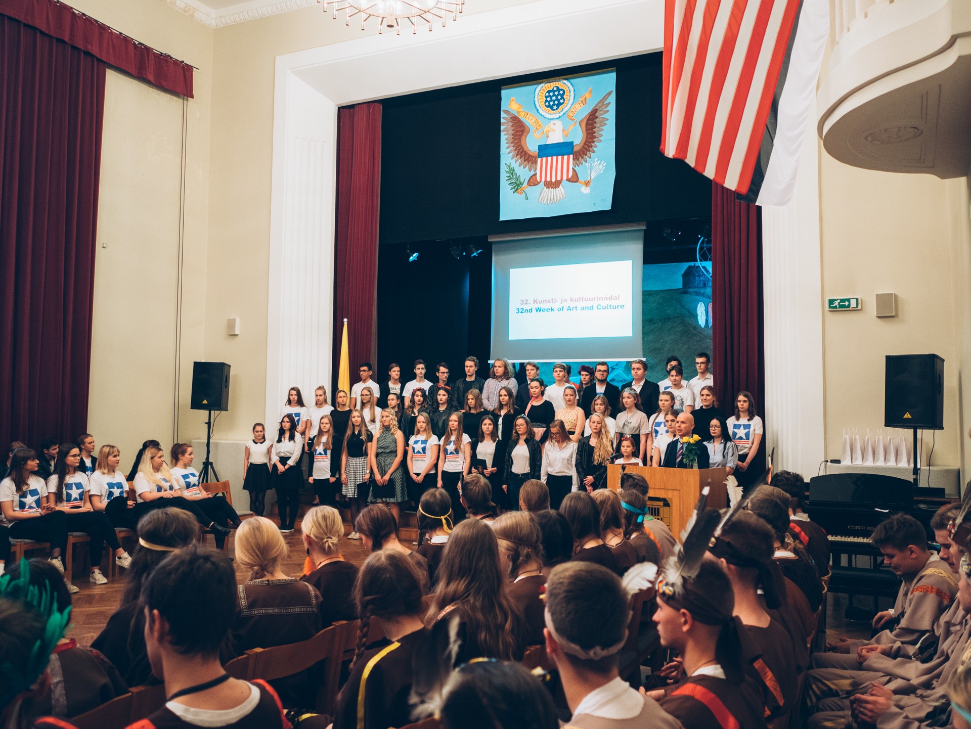 Eine Gruppe von Menschen steht vor einem sitzenden Publikum an einem Podium mit Mikrofonen, flankiert von Flaggen und Lautsprechern, während im Hintergrund ein Bildschirm mit Text und Abbildungen angezeigt wird.