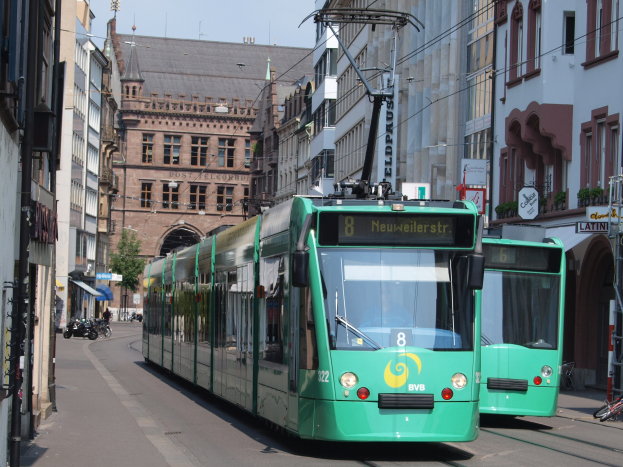 Zwei grüne Straßenbahnen fahren auf einer von hohen Gebäuden gesäumten Straße, mit parkenden Fahrrädern und Fussgängern auf den Gehwegen, einem Baum und einem klaren blauen Himmel im Hintergrund.