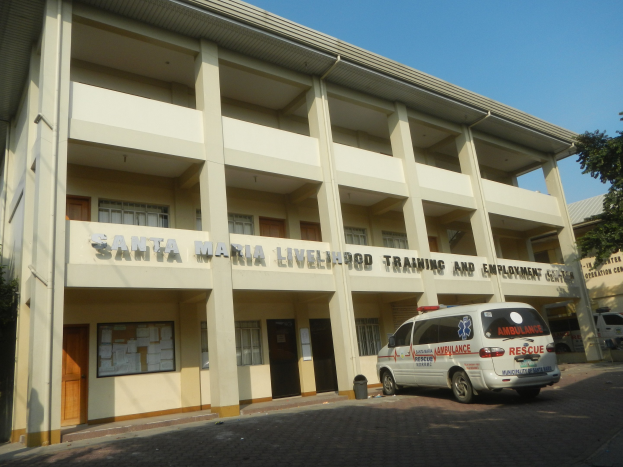 Gebäude des Santa Maria Lebensmittelpunkt- und Arbeitsplatz-Trainingszentrums mit Fenstern, Türen und Beschriftung, umgeben von Bäumen und Fahrzeugen auf der Straße unter einem klaren blauen Himmel.