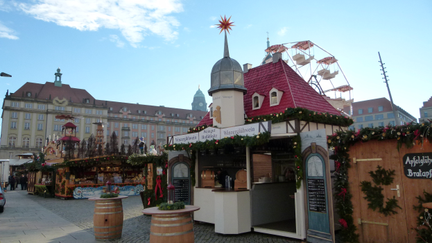 Ein geschäftiger Weihnachtsmarkt in einem Stadtplatz mit geschmückten Bäumen, Gebäuden, Straßenlaternen, Fahrzeugen, Passanten und einem bewölkten Himmel.