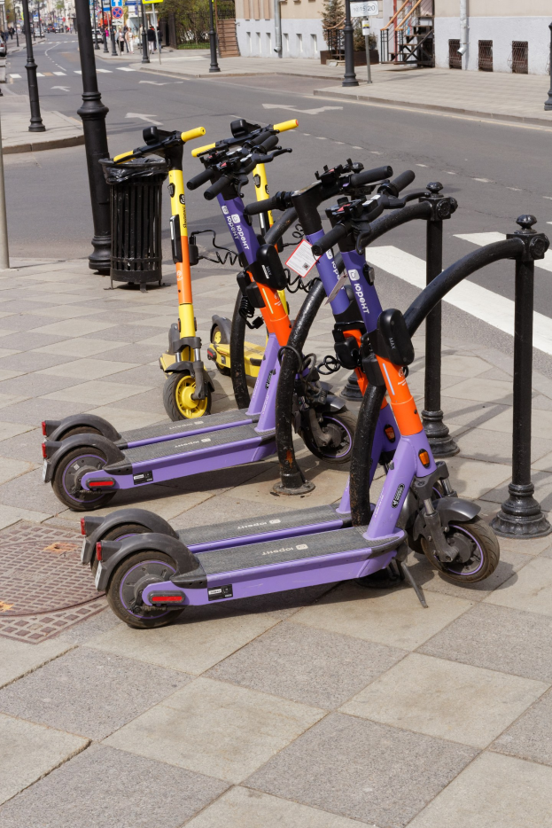 Eine Reihe von Elektro-Scootern, die auf der Straße geparkt sind, mit einem Mülleimer im Vordergrund und Gebäuden, Bäumen und Fahrzeugen im Hintergrund.