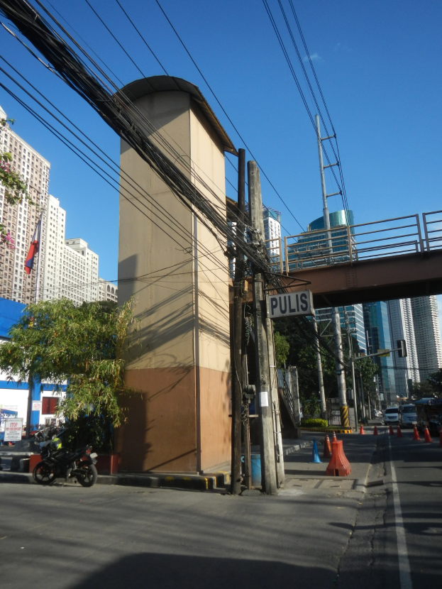 Stadtstraße mit Brücke im Hintergrund, Gebäude, Bäume, Pfähle, Bretter, Verkehrskegel und Fahrzeuge auf der Straße, unter einem sichtbaren Himmel mit Stromleitungen entlang der Straße.
