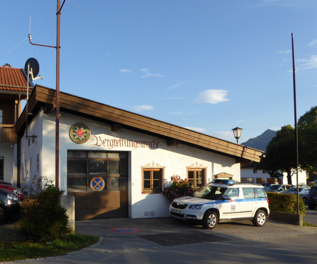 Polizeiauto vor einem Gebäude mit Fenstern und Türen geparkt, umgeben von Grün, Straßenmöbeln und einem bewölkten Himmel.