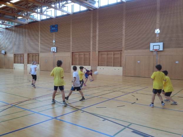 Kinder spielen mit einem Ball vor einer hölzernen Wand mit Basketballkörben, Holzstangen und Fenstern oben.