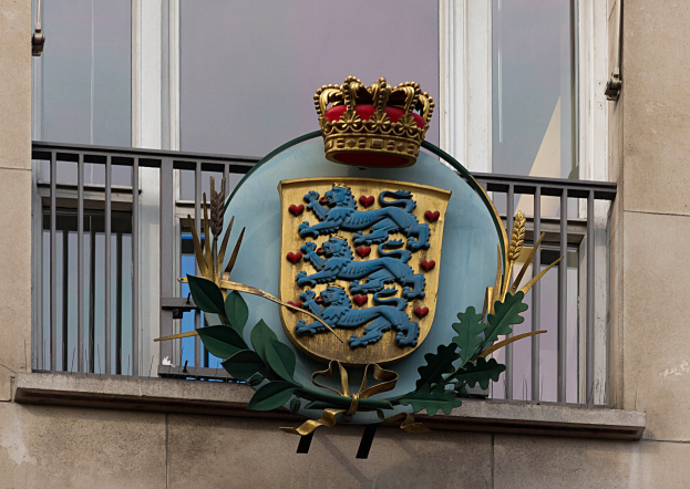 Das britische Königswappen auf der Außenseite eines Gebäudes, mit einer Krone und einem Blatt-Logo, sowie Fenstern und Metallgittern im Hintergrund.