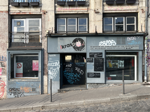 Ein Gebäude mit Fenstern, Geländern, Türen, Texttafeln, Plakaten, Pfosten und Graffiti neben einer Straße.