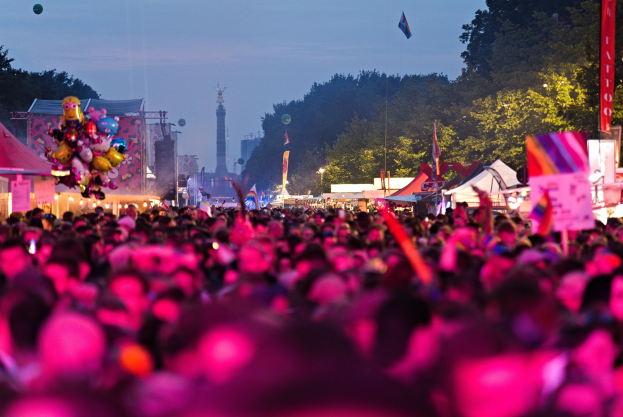 Eine große Menge Menschen bei einem nächtlichen Musikfestival, beleuchtet von farbigen Lichtern, mit Zelten, Fahnen und Pfählen im Vordergrund und Bäumen, einem Turm und einem klaren Himmel im Hintergrund.