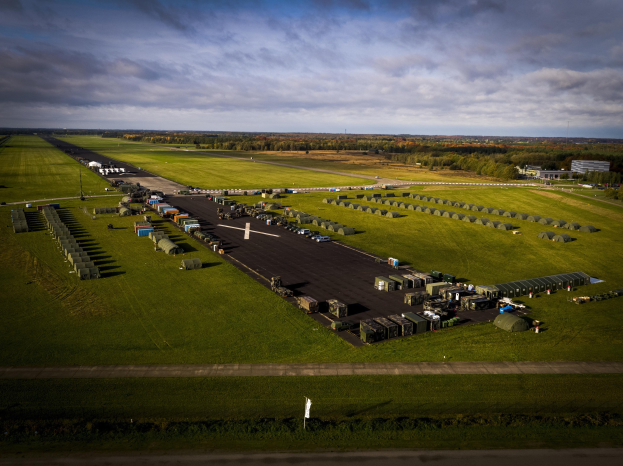 Luftaufnahme einer Militärbasis in den Niederlanden mit Fahrzeugen, Gras, Bäumen, Gebäuden und einem bewölkten Himmel.