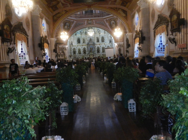 Eine Hochzeitszeremonie in einer Kirche mit Gästen auf Bänken sitzend und im Gang stehend, dekoriert mit Zimmerpflanzen, Skulpturen, Vorhängen und Kronleuchtern, mit einem Bildschirm im Hintergrund.