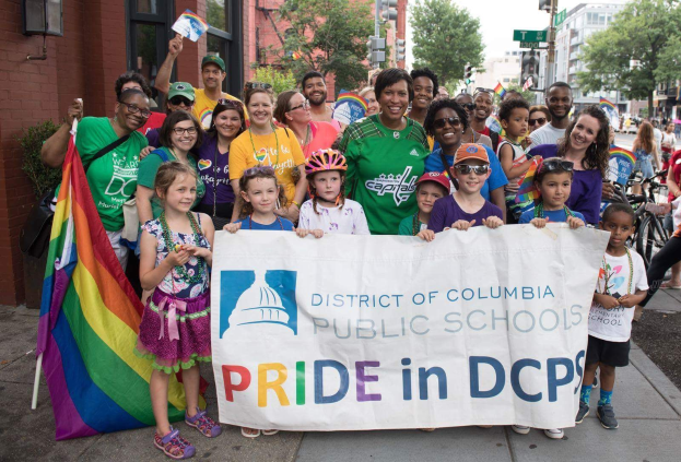 Gruppe von Menschen auf dem Boden stehend, einige halten ein Banner mit der Aufschrift "District of Columbia Public Schools Pride in DCPS", mit Kindern, die eine Fahne halten, einem Radfahrer, Bäumen, Gebäuden, Straßenschildern, Ampeln und einem bewölkten Himmel.